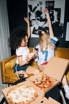 Two young women enjoying a pizza party and red wine indoors, expressing joy.